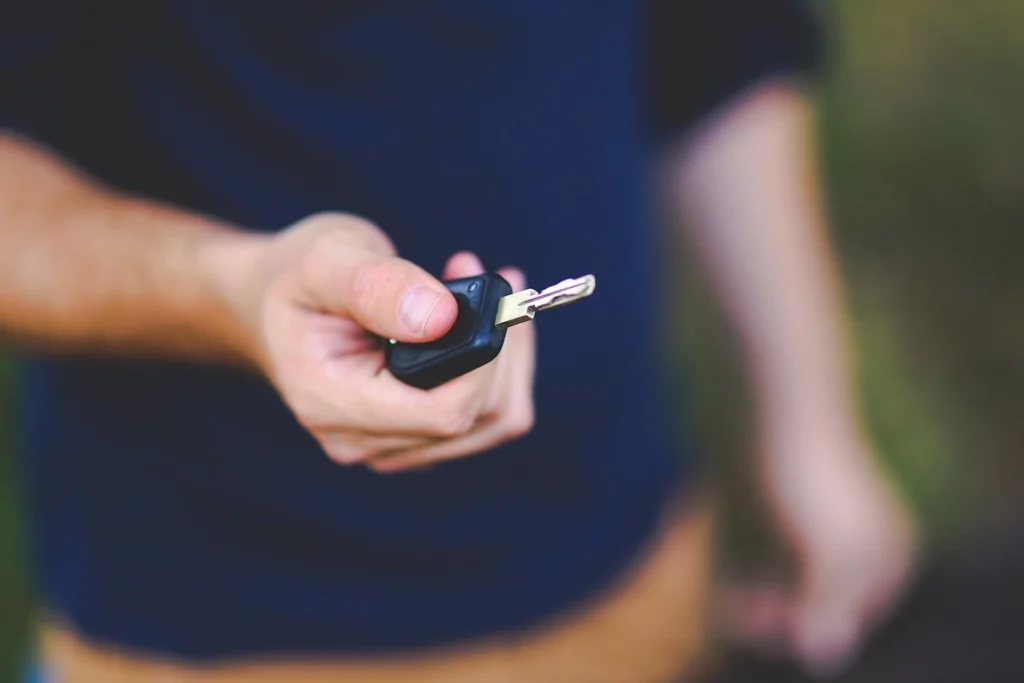 A man with a blue shirt and brown pants is holding out a car key in his right hand.